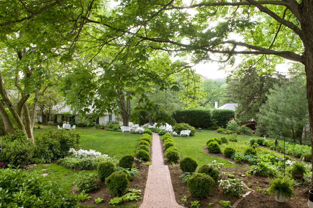 Spring green surrounds the central brick path axis and adjacent serpentine beds edged with boxwoods, hostas and occasional azaleas. An old three-car garage resem-bles an English cottage, gives the back garden a focal point and complements the white of garden furniture and Delaware Valley evergreen azaleas.
